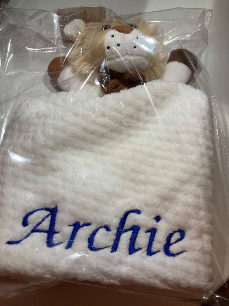 Puede incluir: Una manta de beb&eacute; blanca con el nombre "Archie" bordado en azul. Una cabeza de le&oacute;n de peluche con pelaje marr&oacute;n y beige est&aacute; adjunta. El conjunto est&aacute; empaquetado en pl&aacute;stico transparente.