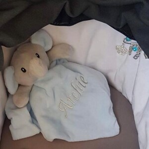 Puede incluir: Un peluche de elefante con una manta azul claro. La manta est&aacute; bordada con el nombre "Archie" en cursiva. El elefante es gris con orejas grandes y una trompa. El juguete est&aacute; sobre una superficie blanca.