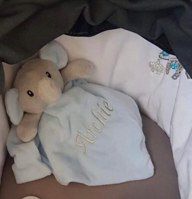 Puede incluir: Un peluche de elefante con una manta azul claro. La manta est&aacute; bordada con el nombre "Archie" en cursiva. El elefante es gris con orejas grandes y una trompa. El juguete est&aacute; sobre una superficie blanca.