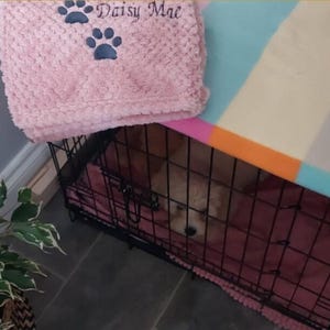 May include: A black metal wire pet crate containing a small light-colored puppy, covered by a pink textured blanket with two dark paw prints and the embroidered text Daisy Mae, and a multi-colored striped fabric top cover.
