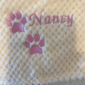 May include: A pale yellow textured fabric blanket featuring two embroidered pink paw prints and the name Nancy stitched in pink cursive script. The material has a soft, dimpled surface pattern throughout the entire piece.