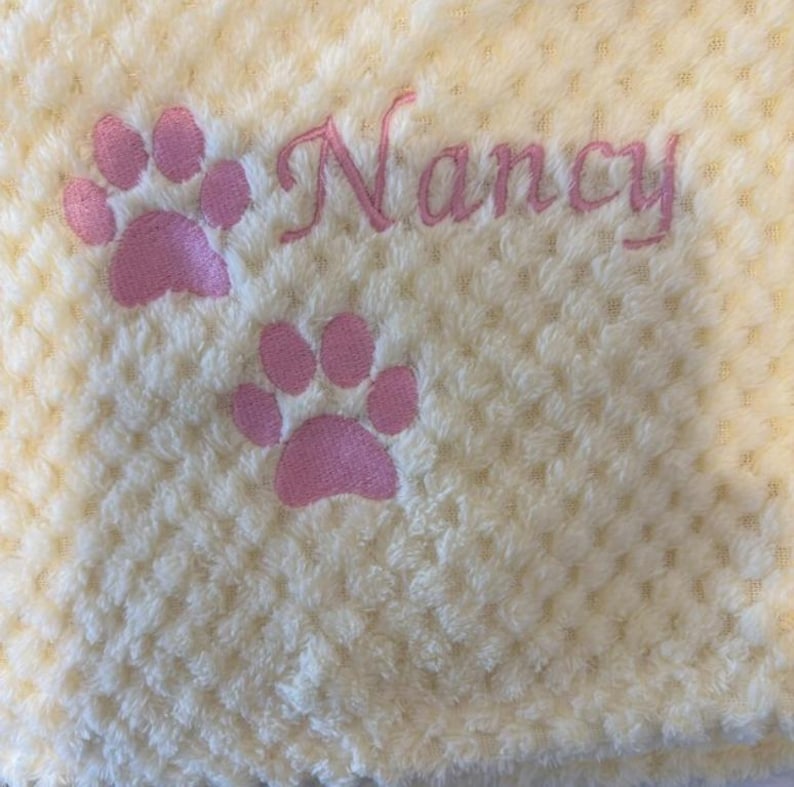 May include: A pale yellow textured fabric blanket featuring two embroidered pink paw prints and the name Nancy stitched in pink cursive script. The material has a soft, dimpled surface pattern throughout the entire piece.