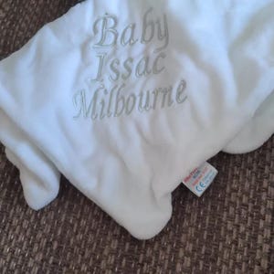 Puede incluir: Una manta blanca para beb&eacute; con el texto bordado "Baby Issac Milbourne" en gris claro. La manta es suave y afelpada, con una etiqueta en la esquina inferior derecha.