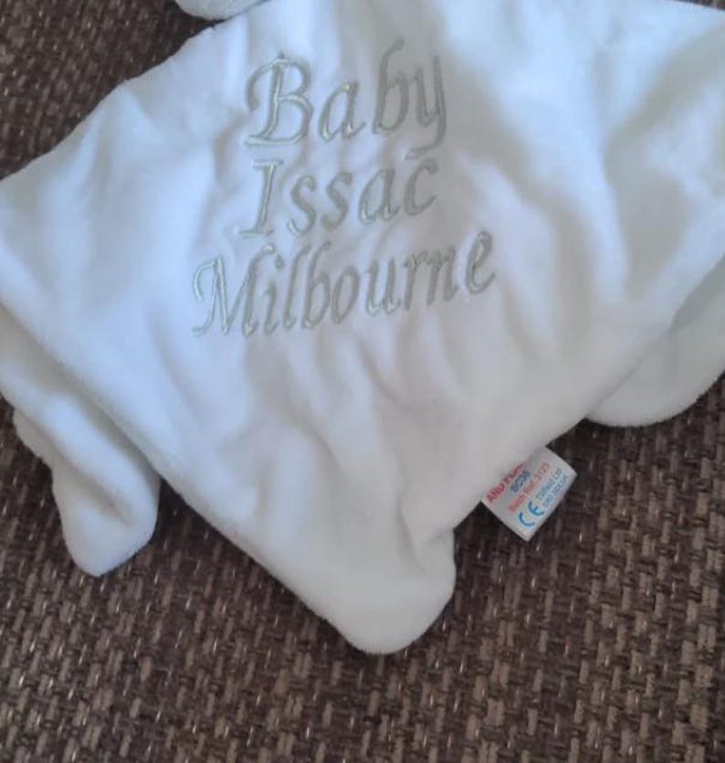 Puede incluir: Una manta blanca para beb&eacute; con el texto bordado "Baby Issac Milbourne" en gris claro. La manta es suave y afelpada, con una etiqueta en la esquina inferior derecha.