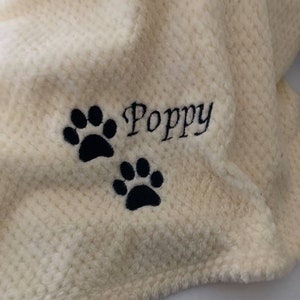 May include: A cream-colored, textured waffle-knit blanket featuring black embroidery. The embroidery consists of the name Poppy written in a cursive script, accompanied by two black paw prints positioned below the name.