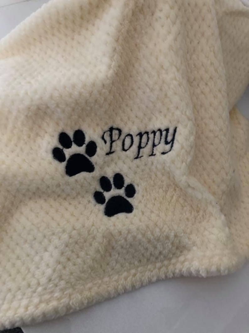 May include: A cream-colored, textured waffle-knit blanket featuring black embroidery. The embroidery consists of the name Poppy written in a cursive script, accompanied by two black paw prints positioned below the name.