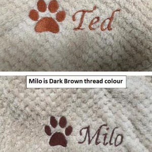 May include: A beige textured fabric featuring two embroidered designs. The top section shows a bronze paw print and the name Ted. The bottom section displays a dark brown paw print and the name Milo, with text labels indicating the thread colors.