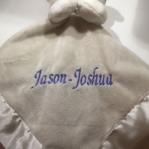 Puede incluir: Una suave manta de seguridad para beb&eacute; de color gris claro con una cabeza de oso de peluche y ribete de sat&eacute;n. La manta presenta el nombre "Jason-Joshua" bordado en hilo azul, lo que la convierte en un regalo personalizado para beb&eacute;s.