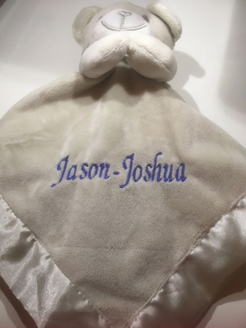 Puede incluir: Una suave manta de seguridad para beb&eacute; de color gris claro con una cabeza de oso de peluche y ribete de sat&eacute;n. La manta presenta el nombre "Jason-Joshua" bordado en hilo azul, lo que la convierte en un regalo personalizado para beb&eacute;s.