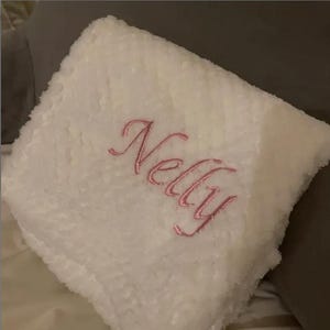 May include: A square white plush pillow with a textured, fuzzy surface. The name Nelly is embroidered in the center in a cursive, pink script font. The pillow has a soft, thick appearance and is folded or positioned at an angle.