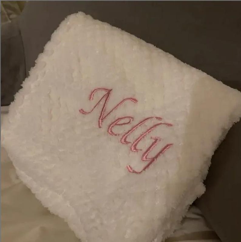 May include: A square white plush pillow with a textured, fuzzy surface. The name Nelly is embroidered in the center in a cursive, pink script font. The pillow has a soft, thick appearance and is folded or positioned at an angle.