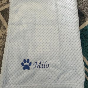 May include: A light blue blanket with a textured waffle pattern. The blanket features an embroidered dark blue paw print icon followed by the name Milo in a cursive script font, positioned in the lower left corner of the fabric.