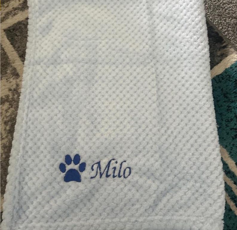 May include: A light blue blanket with a textured waffle pattern. The blanket features an embroidered dark blue paw print icon followed by the name Milo in a cursive script font, positioned in the lower left corner of the fabric.