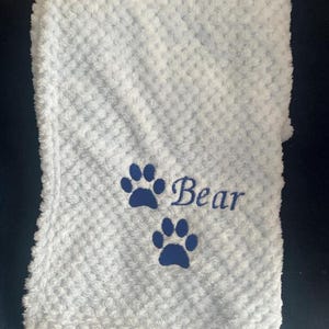 May include: A light blue textured blanket featuring a waffle-like pattern. The fabric is embroidered with two dark blue paw prints and the word "Bear" in a cursive script font, positioned between the two paw prints on the soft material.