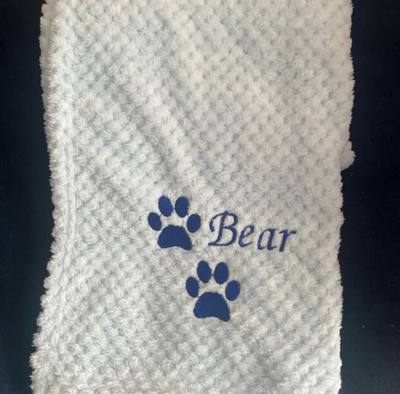 May include: A light blue textured blanket featuring a waffle-like pattern. The fabric is embroidered with two dark blue paw prints and the word "Bear" in a cursive script font, positioned between the two paw prints on the soft material.