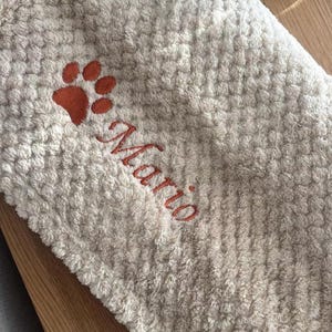 May include: A beige waffle-textured towel featuring an embroidered orange paw print and the name Mario stitched in matching orange thread. The fabric has a soft, raised grid pattern throughout the surface of the textile material.