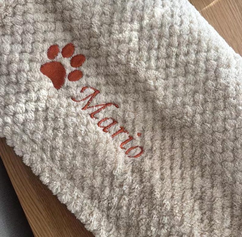 May include: A beige waffle-textured towel featuring an embroidered orange paw print and the name Mario stitched in matching orange thread. The fabric has a soft, raised grid pattern throughout the surface of the textile material.