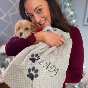 May include: A light gray, textured, plush drawstring bag featuring two dark gray embroidered paw prints and the name LADY embroidered in a dark gray script font on the front surface of the soft fabric material.