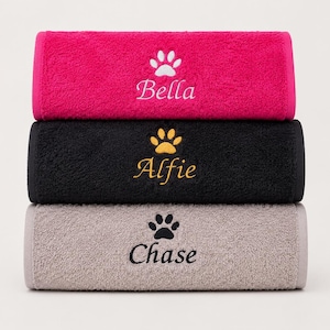 May include: A stack of three folded rectangular towels in hot pink, black, and light gray. Each towel features an embroidered paw print icon above a name: Bella in white, Alfie in gold, and Chase in black, centered on the front surface.