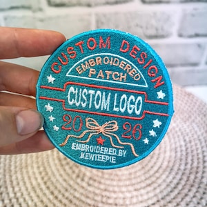 May include: A round, teal embroidered patch with the words "CUSTOM DESIGN" and "CUSTOM LOGO" in red and white. The patch also features the text "EMBROIDERED PATCH" and "2026". The patch is embroidered by "KEWTEEPIE".