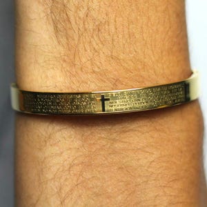 May include: A gold-toned bracelet with a cross and an inscription in Spanish. The bracelet is worn on a person's wrist.