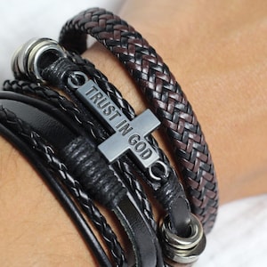 以下が含まれることがあります： 「TRUST IN GOD」と刻まれた銀色の十字架チャームが付いた、重ね付けできる革製のブレスレットセット。セットには、茶色と黒の編み込み革のブレスレットと、複数の黒い革のブレスレットが含まれています。