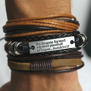 Könnte beinhalten: Ein mehrlagiges Lederarmband mit einem silbernen Anhänger, auf dem "No weapon formed against you shall prosper...Isaiah 54:17" steht.