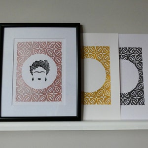 May include: Three framed prints with black frames. The center print features a black and white silhouette of Frida Kahlo with flowers in her hair on a pink background. The print on the left has a gold background and the print on the right has a black background.