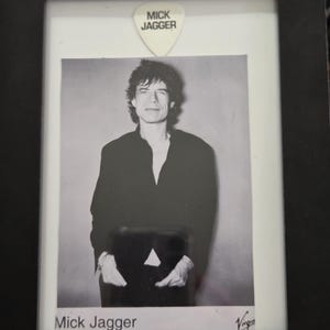 May include: Framed black and white portrait of Mick Jagger. A white guitar pick with "MICK JAGGER" printed on it is above the portrait. The name "Mick Jagger" is printed at the bottom of the portrait.