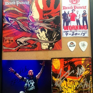 May include: Framed collection of Five Finger Death Punch memorabilia. Includes a CD cover with a skull design, a promotional photo, guitar picks, and a signed photo of a band member. The color scheme is primarily red, black, and white.