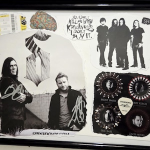 May include: A framed collage featuring Shinedown band memorabilia, including a signed photograph of the four members, a concert ticket, a guitar pick, and a JSA certification card. The display includes text such as "Don't be delicate, be vast and brilliant" and "You can't kill me with kindness, I don't buy it."