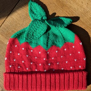 K&ouml;nnte beinhalten: Gestrickte Erdbeerm&uuml;tze mit rotem K&ouml;rper und gr&uuml;nen Bl&auml;ttern. Die M&uuml;tze hat wei&szlig;e Samen Details und einen gerippten Saum. Ein lustiges und originelles Modeaccessoire.