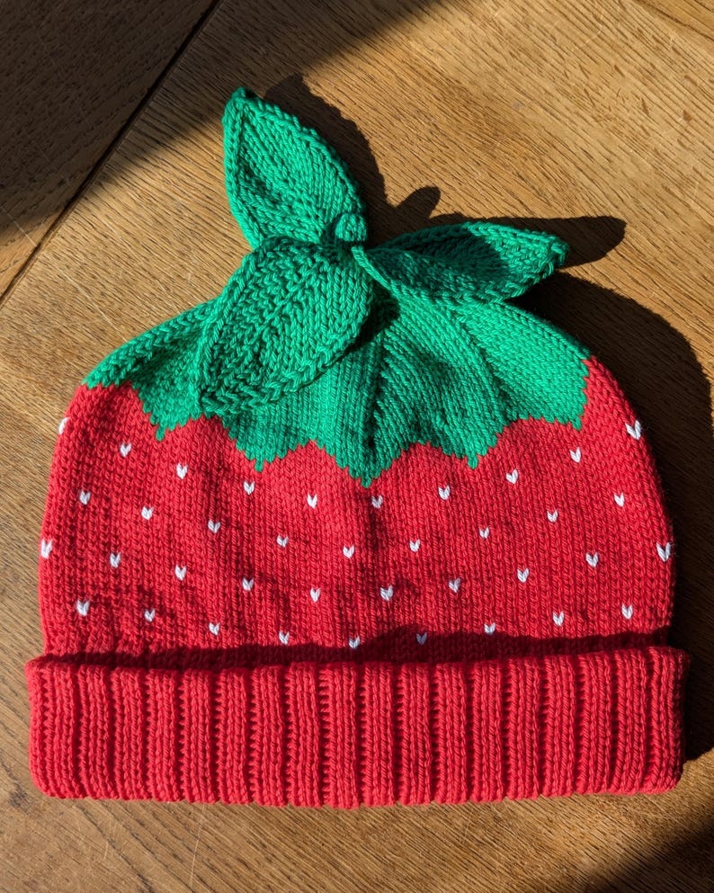 K&ouml;nnte beinhalten: Gestrickte Erdbeerm&uuml;tze mit rotem K&ouml;rper und gr&uuml;nen Bl&auml;ttern. Die M&uuml;tze hat wei&szlig;e Samen Details und einen gerippten Saum. Ein lustiges und originelles Modeaccessoire.