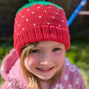 K&ouml;nnte beinhalten: Eine rote Strickm&uuml;tze mit einem gr&uuml;nen Stiel, die einer Erdbeere nachempfunden ist. Die M&uuml;tze hat kleine wei&szlig;e herzf&ouml;rmige Details. Die M&uuml;tze wird von einer Person getragen, die eine rosa Kapuzenjacke tr&auml;gt.