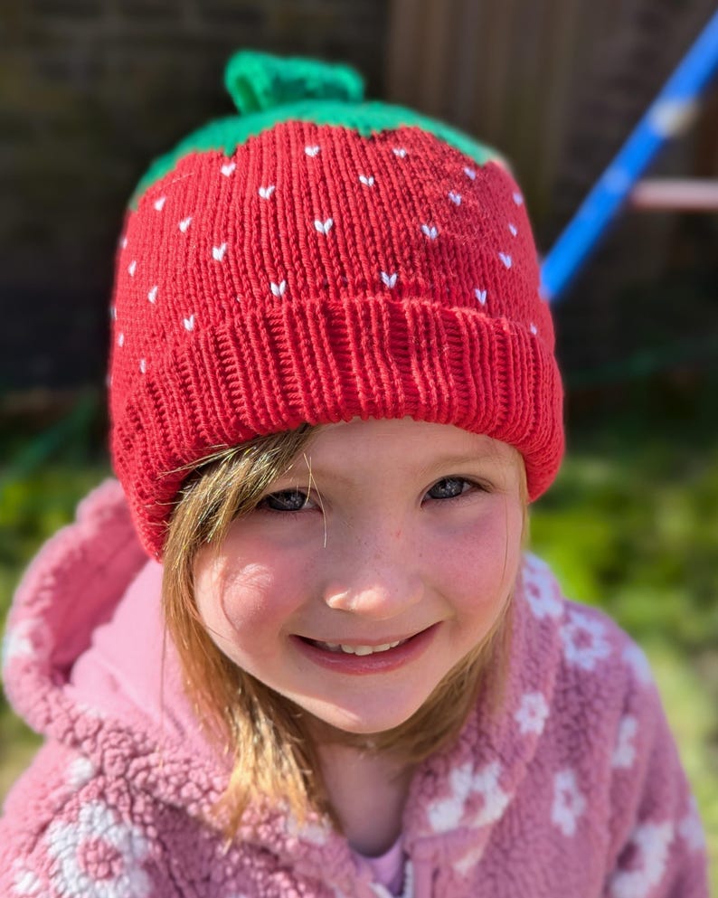 K&ouml;nnte beinhalten: Eine rote Strickm&uuml;tze mit einem gr&uuml;nen Stiel, die einer Erdbeere nachempfunden ist. Die M&uuml;tze hat kleine wei&szlig;e herzf&ouml;rmige Details. Die M&uuml;tze wird von einer Person getragen, die eine rosa Kapuzenjacke tr&auml;gt.