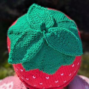 K&ouml;nnte beinhalten: Gestrickte Erdbeerm&uuml;tze. Die M&uuml;tze ist rot mit wei&szlig;en Samendetails und einem gr&uuml;nen Blattmuster. Ein lustiges Accessoire f&uuml;r Kinder, aus Wolle gefertigt.
