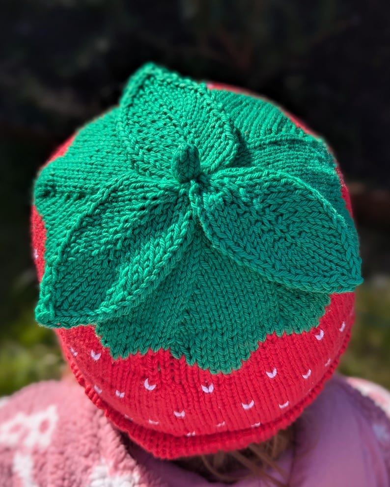 K&ouml;nnte beinhalten: Gestrickte Erdbeerm&uuml;tze. Die M&uuml;tze ist rot mit wei&szlig;en Samendetails und einem gr&uuml;nen Blattmuster. Ein lustiges Accessoire f&uuml;r Kinder, aus Wolle gefertigt.