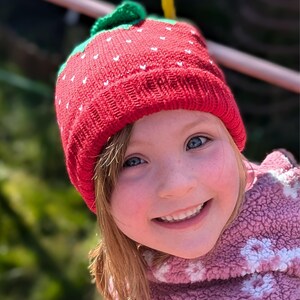 K&ouml;nnte beinhalten: Eine rote Strickm&uuml;tze mit gr&uuml;nen Bl&auml;ttern und wei&szlig;en Samen, die einer Erdbeere &auml;hnelt. Die M&uuml;tze wird von einer Person mit heller Haut und blauen Augen getragen, die l&auml;chelt. Die Person tr&auml;gt eine rosa Fleecejacke.