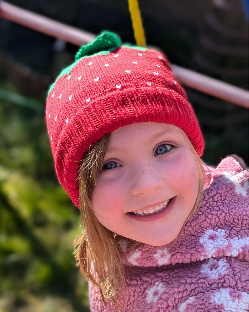 K&ouml;nnte beinhalten: Eine rote Strickm&uuml;tze mit gr&uuml;nen Bl&auml;ttern und wei&szlig;en Samen, die einer Erdbeere &auml;hnelt. Die M&uuml;tze wird von einer Person mit heller Haut und blauen Augen getragen, die l&auml;chelt. Die Person tr&auml;gt eine rosa Fleecejacke.