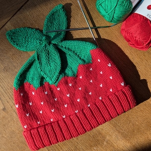 K&ouml;nnte beinhalten: Gestrickte Erdbeerm&uuml;tze mit gr&uuml;nem Blattdesign. Die M&uuml;tze ist rot mit wei&szlig;en Samendetails und einem umgeschlagenen Saum. Stricknadeln und Kn&auml;uel aus gr&uuml;ner und roter Wolle befinden sich im Hintergrund.