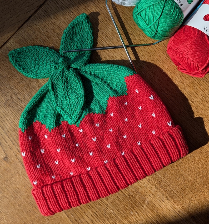 K&ouml;nnte beinhalten: Gestrickte Erdbeerm&uuml;tze mit gr&uuml;nem Blattdesign. Die M&uuml;tze ist rot mit wei&szlig;en Samendetails und einem umgeschlagenen Saum. Stricknadeln und Kn&auml;uel aus gr&uuml;ner und roter Wolle befinden sich im Hintergrund.