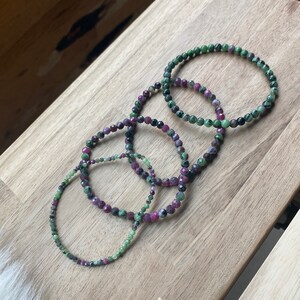May include: Four green and pink gemstone bracelets stacked on a wooden surface. The bracelets are made of small, round beads.