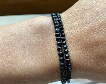 May include: Two black beaded bracelets stacked on a person's wrist. The beads are small and round, and the bracelets are simple and elegant.