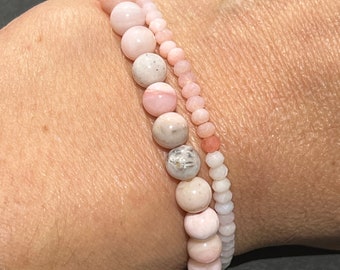 Könnte beinhalten: Zwei Armbänder aus rosa und weißen Opalperlen. Die Armbänder sind auf dem Handgelenk einer Person übereinander gelegt.