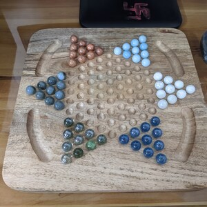 May include: A wooden game board with a grid of holes and five sets of marbles in different colors: blue, green, white, gray, and brown. The marbles are arranged in a star shape.