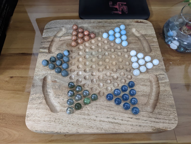 May include: A wooden game board with a grid of holes and five sets of marbles in different colors: blue, green, white, gray, and brown. The marbles are arranged in a star shape.