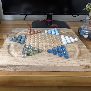 May include: A wooden game board with a star-shaped grid and marble-like pieces in blue, green, and brown colors. The board is made of light brown wood and has a smooth surface.