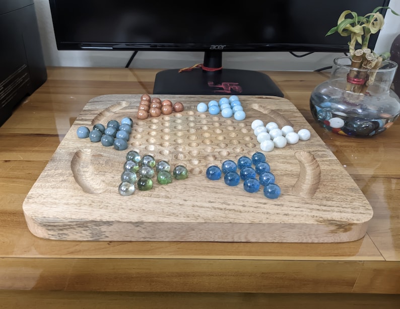 May include: A wooden game board with a star-shaped grid and marble-like pieces in blue, green, and brown colors. The board is made of light brown wood and has a smooth surface.