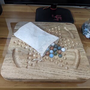 May include: A wooden game board with a white cloth bag filled with colorful marbles. The board has a large number of holes and two curved grooves.