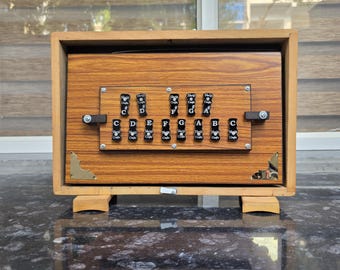 May include: A wooden harmonium instrument featuring a rectangular frame with a light brown wood grain finish. The front panel displays two rows of black keys labeled with musical notes C, D, E, F, G, A, B, and C, with metallic corner accents.